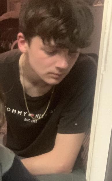A young man with dark hair looks down and to the right, wearing a black t-shirt with a gold chain.