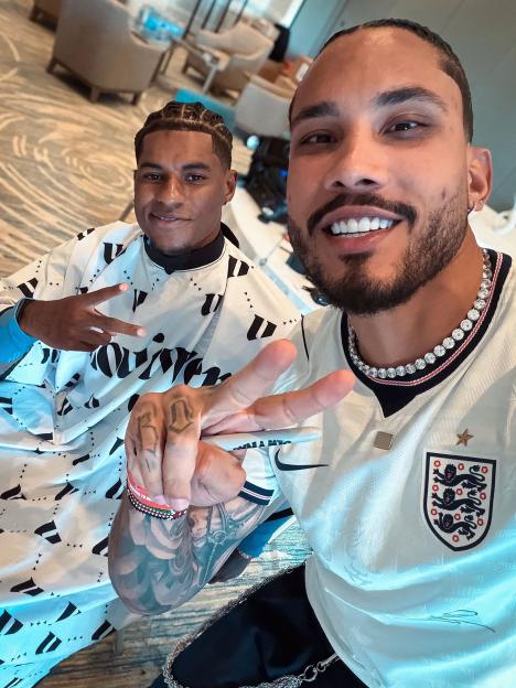 Marcus Rashford sitting in a barber's cape, posing for a photo with his barber who is wearing an England football jersey.