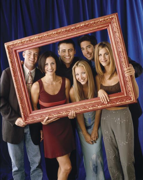 The cast of Friends pose against a blue backdrop, holding a large gold picture frame, with Matthew Perry and Courteney Cox on the left, Matt LeBlanc, Lisa Kudrow, and David Schwimmer in the center, and Jennifer Aniston on the right.