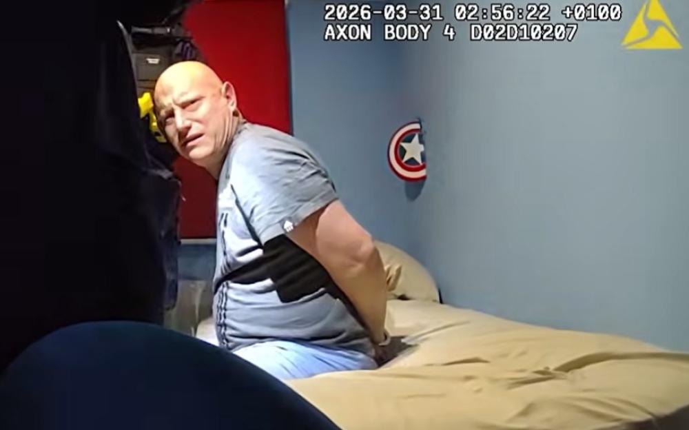 A man in a grey t-shirt and light blue jeans sits on a bed with his hands cuffed behind his back, looking towards the camera.
