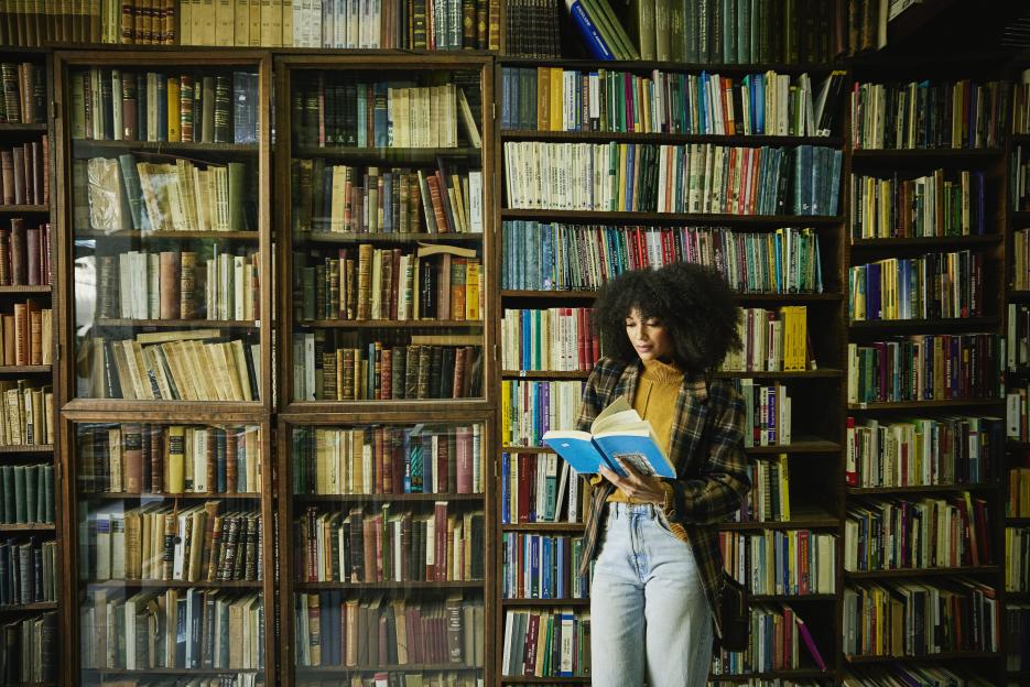 A woman with curly hair stands in an antique bookstore, reading a blue book, surrounded by towering bookshelves filled with various books.