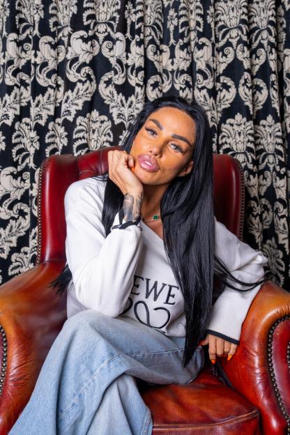 Katie Price sitting in a brown leather armchair, leaning her chin on her hand, against a black and white damask curtain.