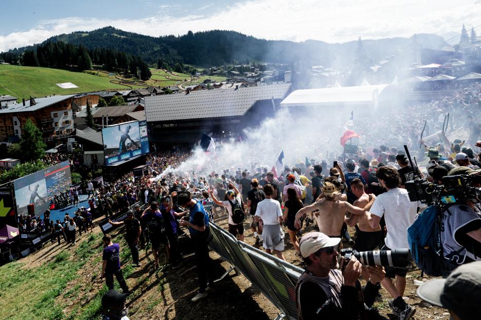 Crowds of spectators cheer and wave flags and smoke flares at the UCI Mountain Bike World Series Haute Savoie 2026.