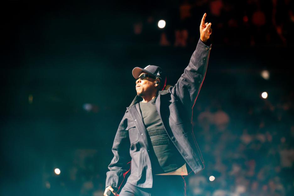 Jay-Z performing on stage, wearing a dark jacket, hat, and sunglasses, with one arm raised.