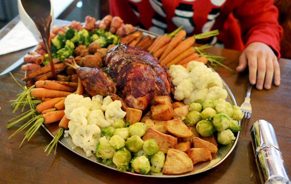 A large holiday meal being served, with gravy being poured onto a platter of roasted meats, carrots, cauliflower, Brussels sprouts, potatoes, and other vegetables.