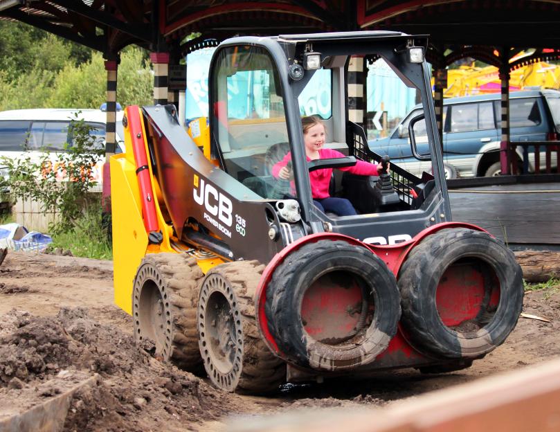 A girl in a pink shirt operating a yellow and black JCB skid steer on a muddy track.