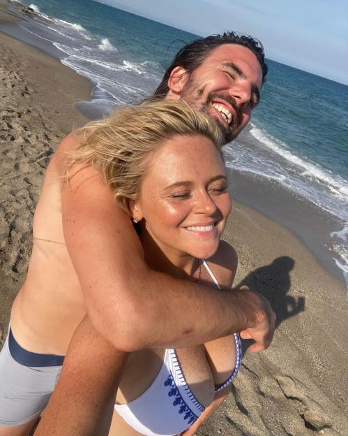 Emily Atack smiling while being hugged from behind by a man on a beach.