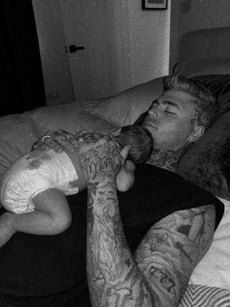 Black and white photo of a tattooed man lying down with a baby sleeping on his chest.