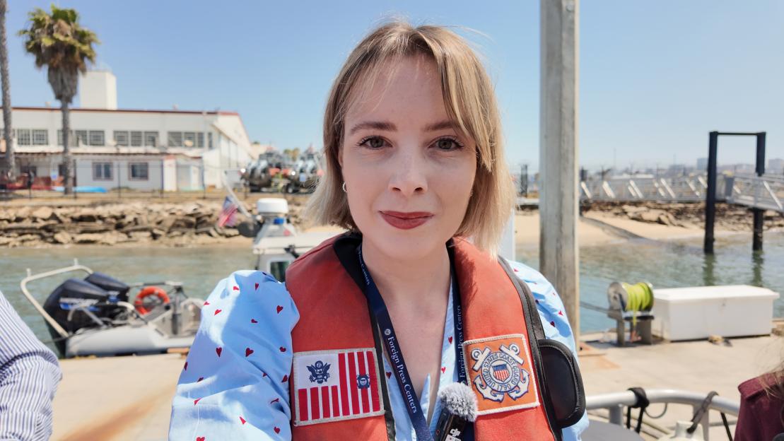 A woman in a life vest with US Coast Guard patches stands on a boat with a harbor and buildings in the background.