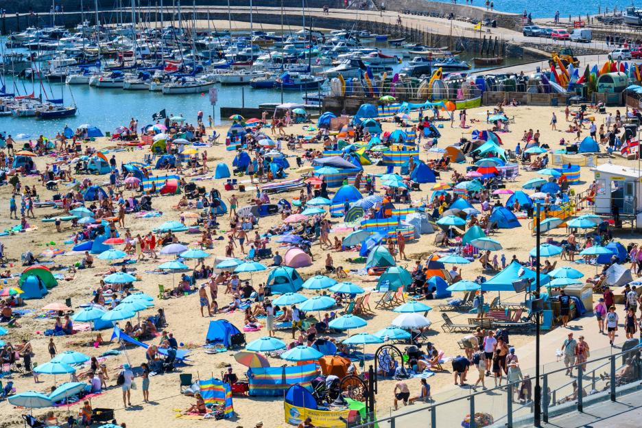 A packed beach with many people, tents, and umbrellas under a bright sun, next to a harbor filled with boats.