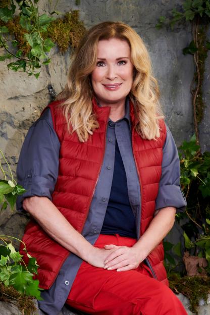 Beverley Callard, wearing red trousers and a red vest over a gray shirt, sits on a stone wall.