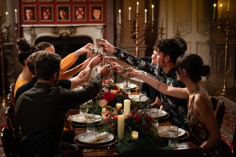 The Cast of The Traitors S4 raising their goblets in a toast around a long dining table.