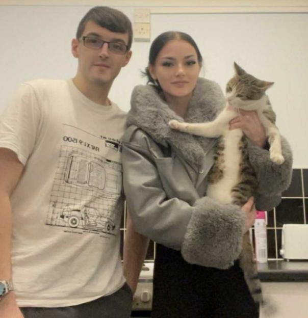 A man in a white t-shirt and glasses stands next to a woman in a grey coat holding a cat.