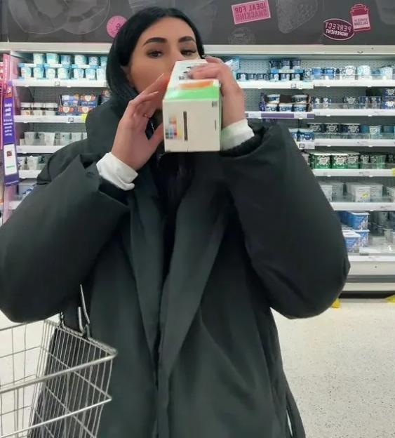 A woman holding a box of goods to her face while standing in a grocery store aisle.
