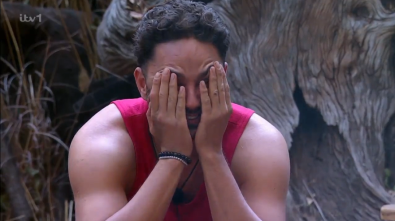 A man in a pink tank top covers his eyes with his hands.
