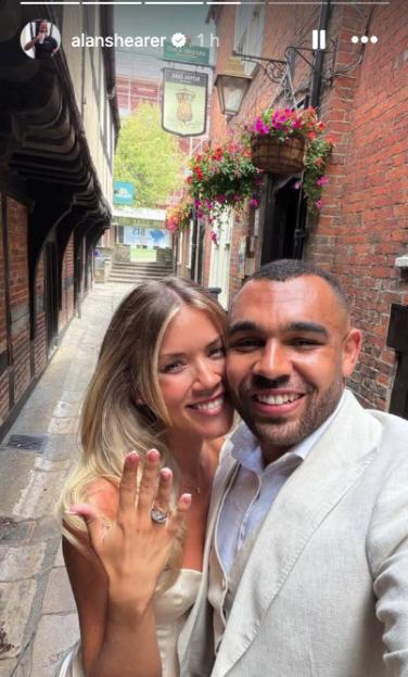 A smiling couple, the woman showing off her engagement ring, in an alleyway decorated with hanging flower baskets.