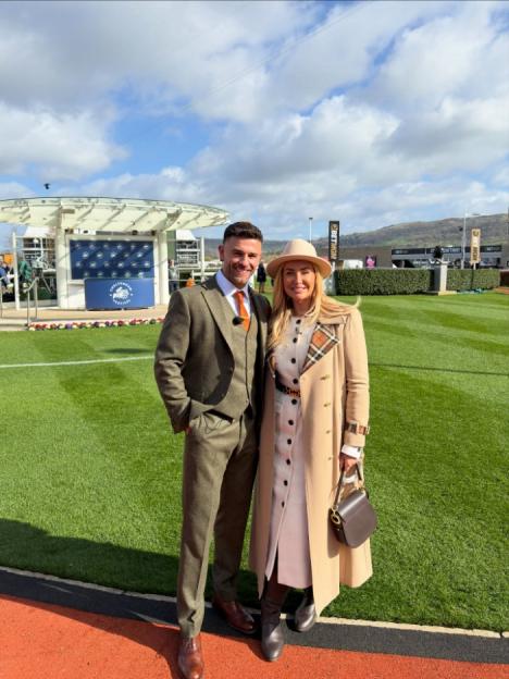 A man in a green tweed suit and an orange tie and a woman in a tan coat and matching hat smile at the camera while standing on a grassy field.