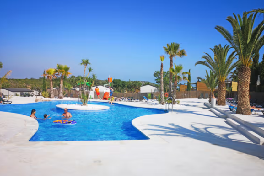 Orangeraie campsite in Càlig, Costa Del Azahar, Spain, with people enjoying a swimming pool and palm trees under a clear blue sky.