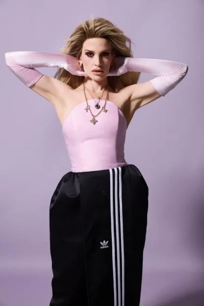 Dakota Schiffer posing in a pink top, black Adidas trackpants, and pink opera gloves against a purple background.