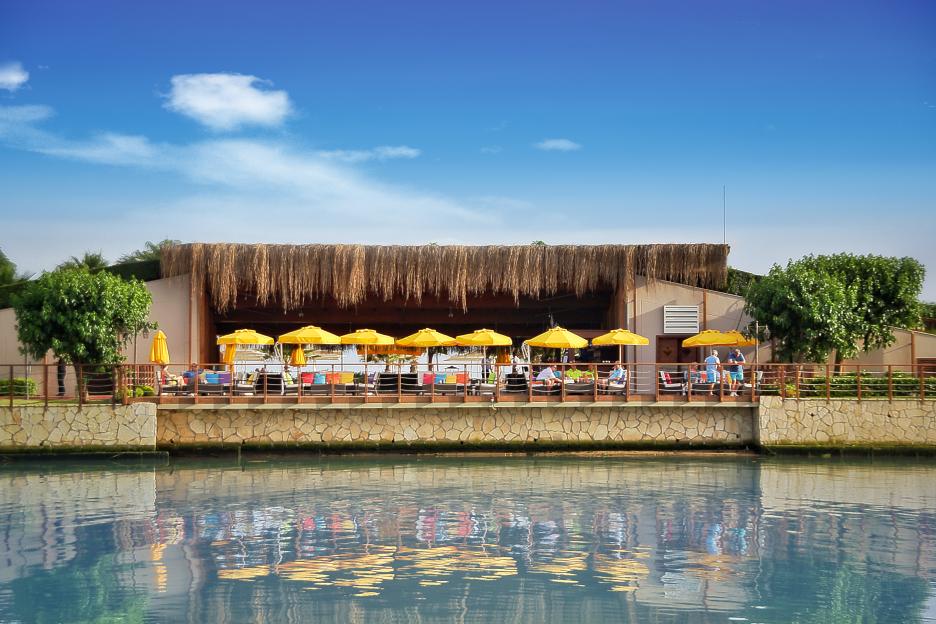 A resort building with a thatched roof and yellow umbrellas, reflecting on the water.