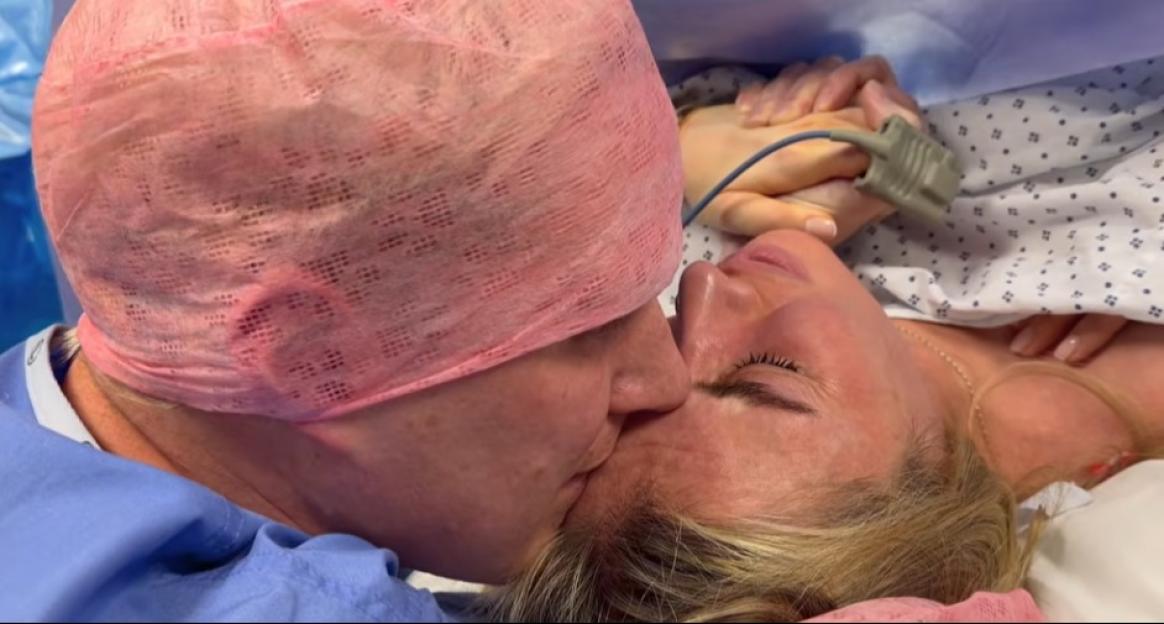 A man in a surgical cap kissing a woman on the forehead who is lying down with a pulse oximeter on her finger.