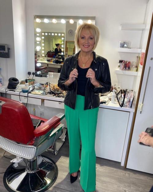 Sue Cleaver in a black leather jacket and green pants, smiling in a dressing room.