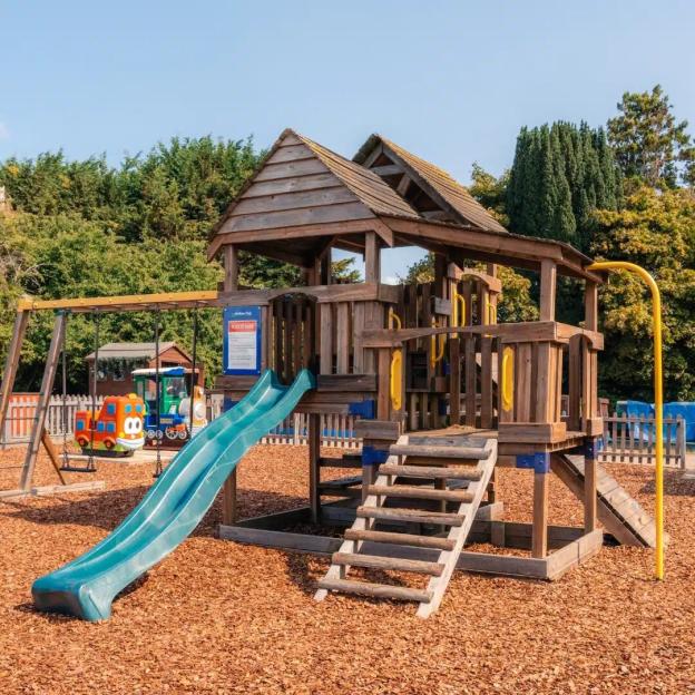 A wooden playground structure with a blue slide, swing set, and a train-shaped spring rider on a wood chip ground.