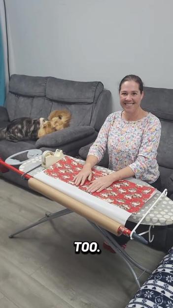 A woman sitting at an ironing board with two cats and gift wrap.