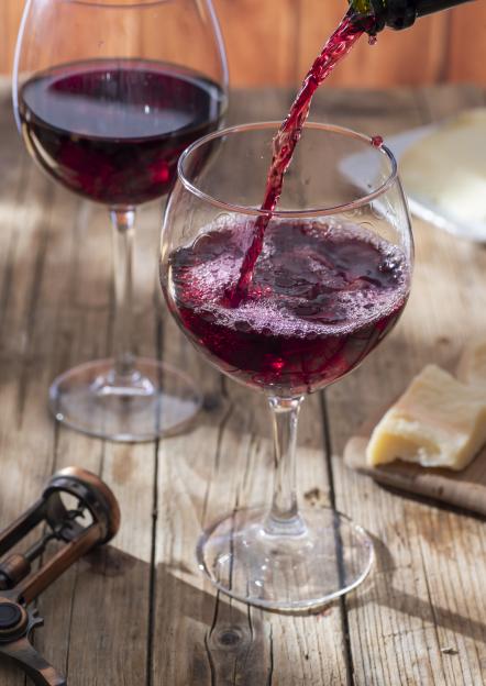 Red wine being poured into a goblet glass, next to another full glass of wine, with cheese and a corkscrew on a wooden table.