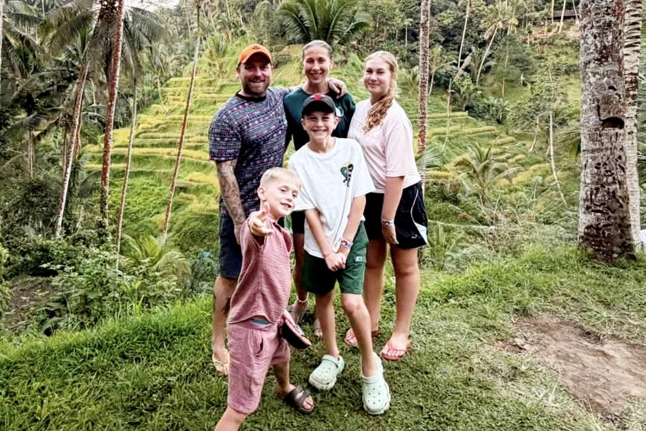 Family of five smiling in Bali with rice paddies and palm trees in the background.