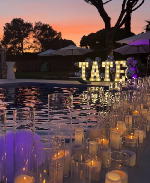 Outdoor evening party setup with "TATE" in marquee letters by a swimming pool lined with candles in glass cylinders.