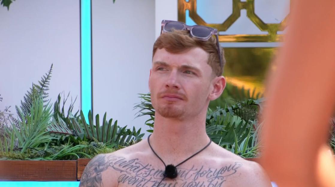 A man with red hair and a beard, wearing sunglasses on his head, a black necklace, and chest tattoos, looks to the left.