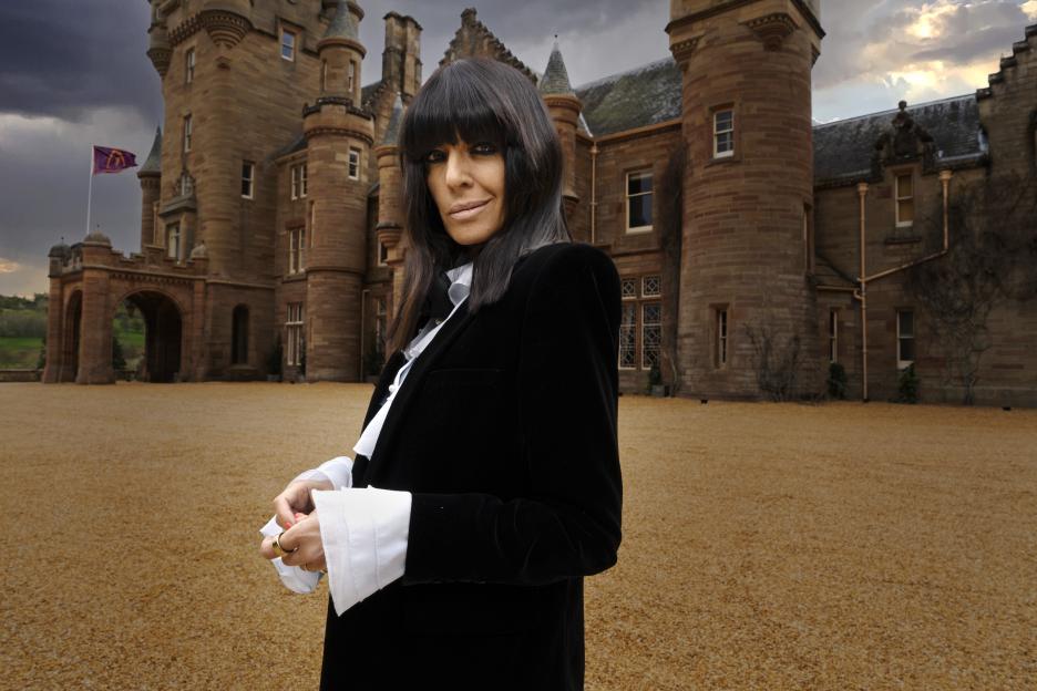 Claudia Winkleman standing in front of a castle, wearing a black velvet jacket and white ruffled shirt.