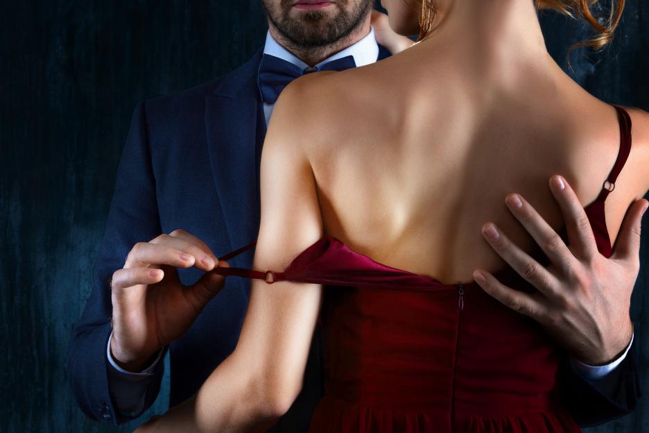 A man in a suit untying a woman's red dress strap while embracing her.