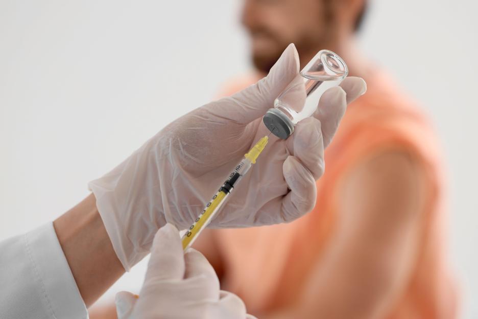 Close-up of a doctor's gloved hands drawing vaccine into a syringe, with a patient's shoulder visible in the background.