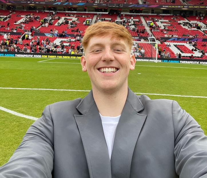 A man with red hair in a gray suit jacket and white t-shirt takes a selfie on a soccer field in front of a stadium with red seats filled with spectators.