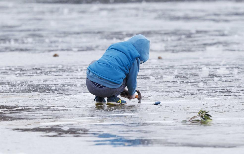 A child in a blue hooded jacket and gray sweatpants kneels on a frozen pond, hitting the ice with a hammer.