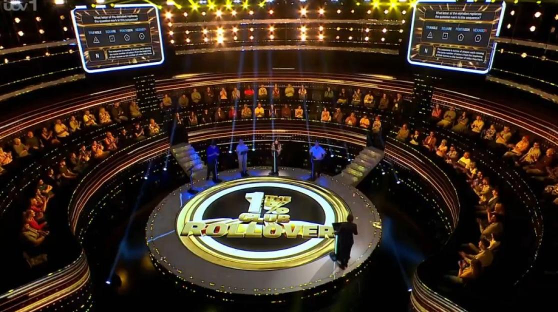A game show set with a large audience surrounding a central stage, where a question is displayed on two screens.