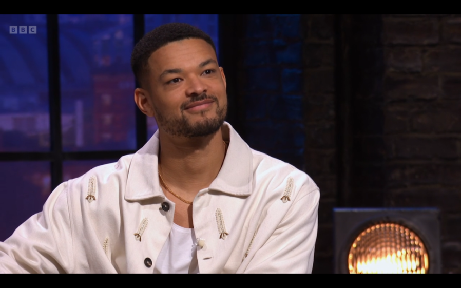 A man with a beard, wearing a cream jacket with embroidered details, smiles slightly while looking to the right.