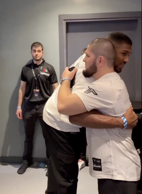 Two men in white shirts embracing, with a man in a black polo shirt standing behind them.