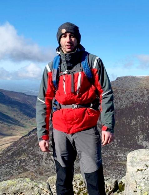 Kristian Hudson in red and gray hiking gear and a black beanie, standing on a rocky mountain with a valley in the background.