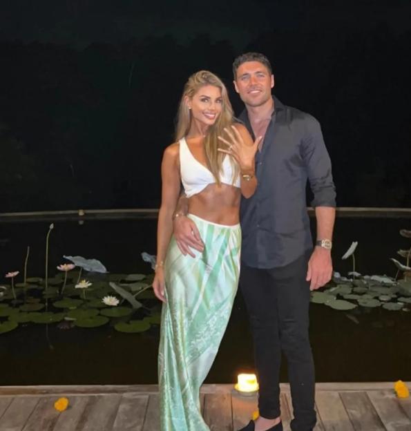 Tom Pearce and his fiancee on a wooden deck next to a pond at night, with the woman showing off her engagement ring.