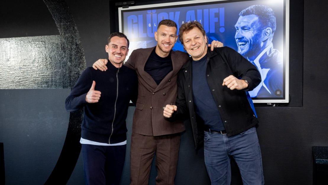 Three men posing for a photo, with a large screen behind them displaying a man's face and text.