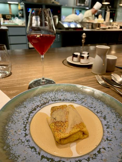 A dish of omelet with sauce on a decorative plate, next to a glass of red wine, with a chef in the background.