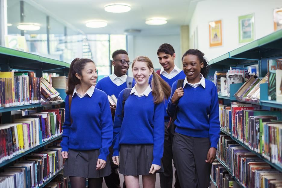 Smiling students wearing blue school uniforms walking between bookshelves in library