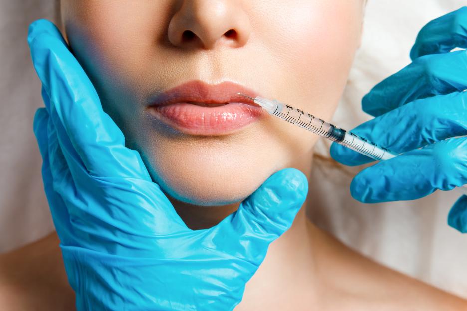 A woman receiving a lip injection, with gloved hands holding a syringe to her lips.