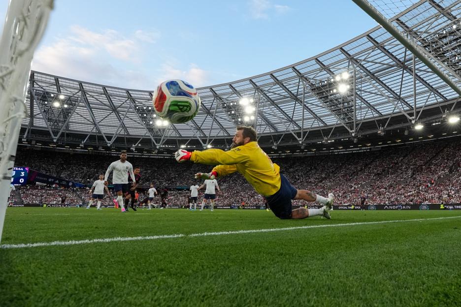 A soccer goalkeeper dives to block a shot during a match at London Stadium.