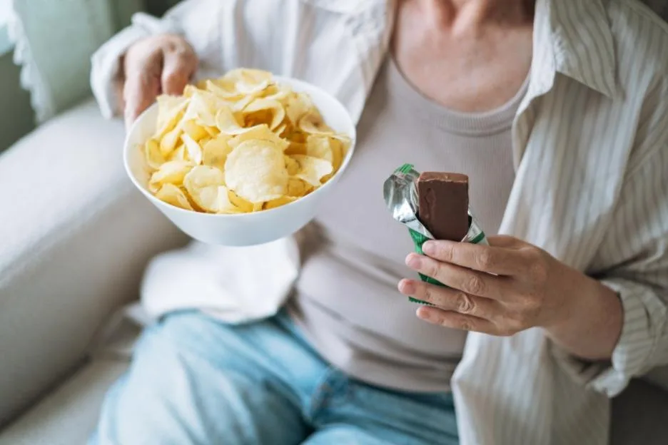 Middle-aged woman sitting on a couch holding a plate of potato chips and a chocolate bar, depicting an unhealthy lifestyle, emotional eating, and an unfulfilled diet