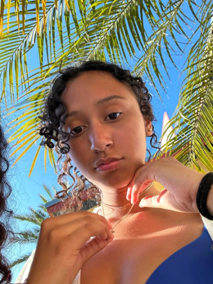 A young woman with curly hair looks at the camera from below, framed by palm tree fronds and a clear blue sky.