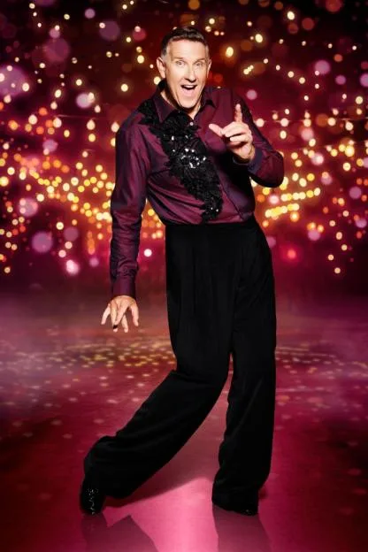 Graeme Hall posing in a sequined purple shirt and black trousers against a festive backdrop.